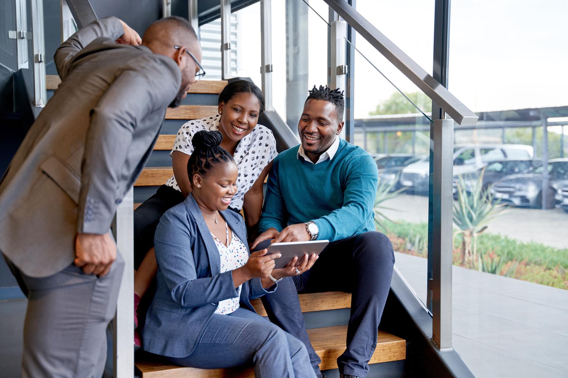 Quatre collègues masculins et féminins professionnels s'asseyant sur l'escalier souriant pratiquant le travail d'équipe sur la tablette d'entreprise